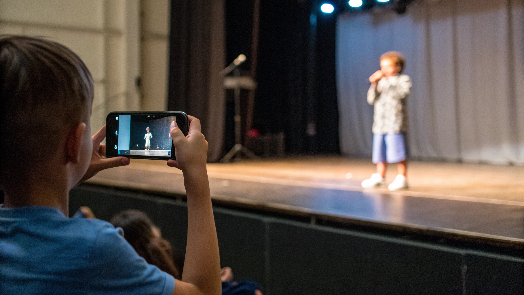 Пользователь снимает ребёнка на сцене на iPhone с оптическим зумом