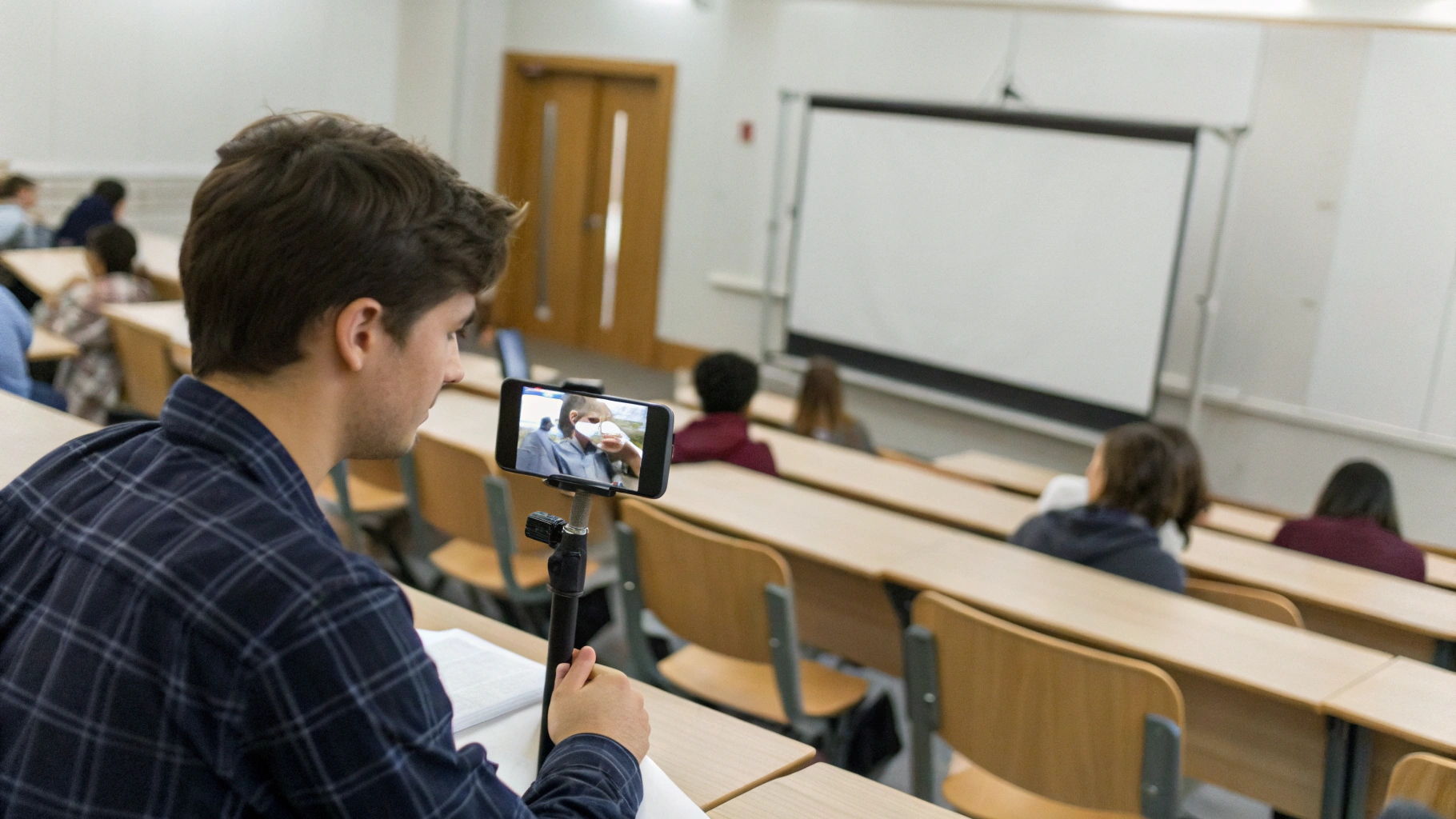 Студент записывает лекцию в аудитории на смартфон, лежащий на парте